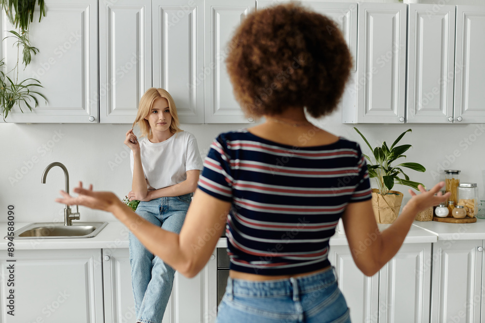 Two women in a white kitchen, one blonde sitting and the other with curly brown hair standing with hands raised Two women in a white kitchen, one blonde sitting and the other with curly brown hair standing with hands raised