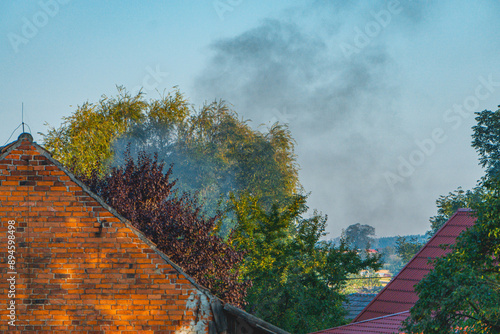 Pożar budynku na wsi, gęsty dym podczas pożaru unoszący się nad domem w centrum wsi. gorące lato, zagrożenie pożarowe photo