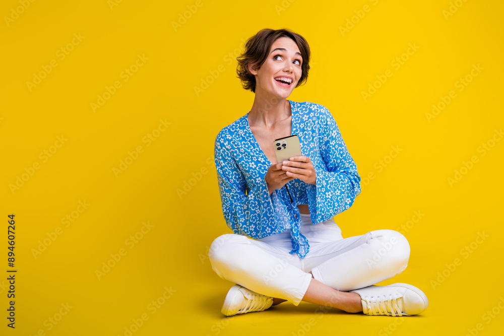Full length photo of impressed girl dressed print blouse sit look at offer empty space hold smartphone isolated on yellow color background Full length photo of impressed girl dressed print blouse sit look at offer empty space hold smartphone isolated on yellow color background