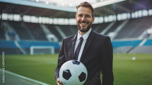 A bearded man in a suit and tie, holding a soccer ball, smiles confidently in an empty stadium, embodying leadership and passion for sports. photo