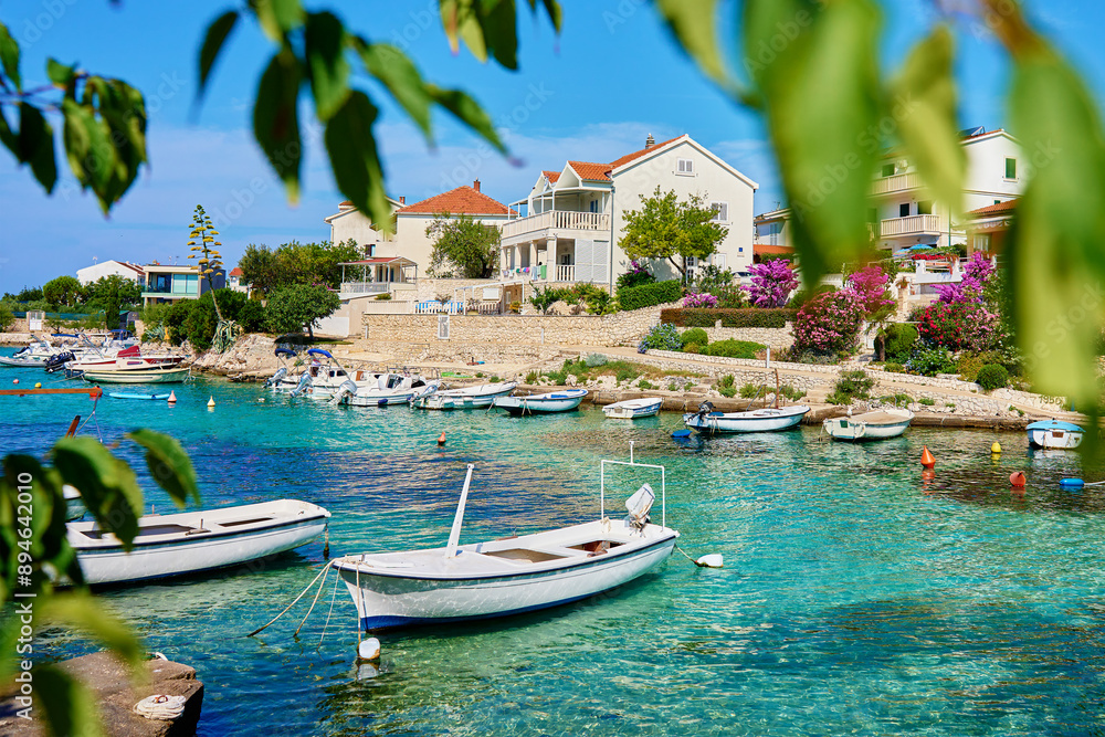 Fototapeta premium Small motorboats are floating on turquoise water near a stone pier by row of beautiful houses with lush gardens. Summer vacation on Adriatic Sea in Rogoznica, Croatia