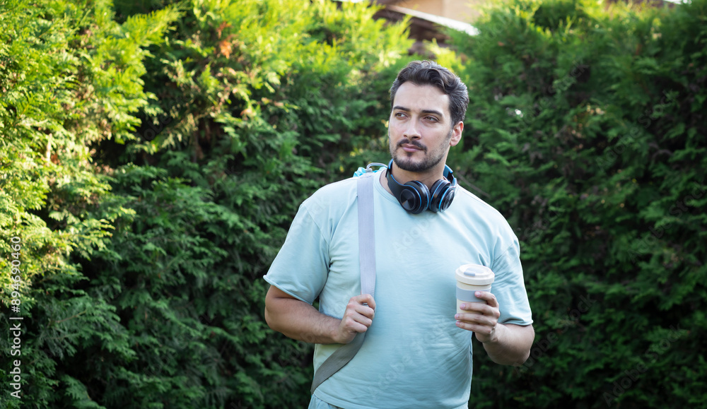 Serious and handsome bearded man in headphones holding takeaway coffee and going to yoga Serious and handsome bearded man in headphones holding takeaway coffee and going to yoga