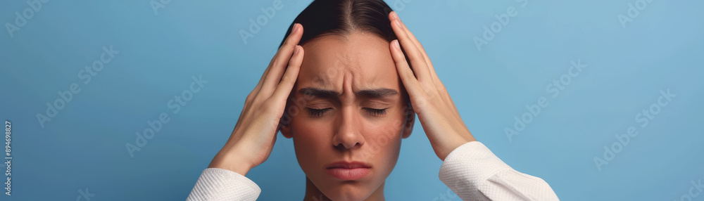 Naklejka premium Young woman holding her head in frustration against a blue background, depicting stress, anxiety, or headache.