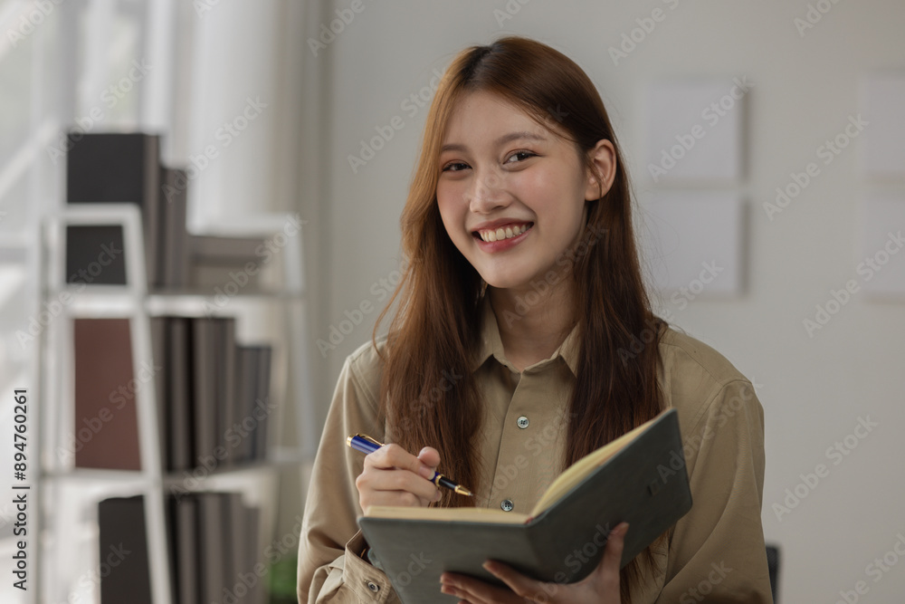 an Asian businesswoman is writing detail from the last meeting, there is a beautiful employee is smiling in the workplace, taking a note into the book witch is the first draft of report an Asian businesswoman is writing detail from the last meeting, there is a beautiful employee is smiling in the workplace, taking a note into the book witch is the first draft of report