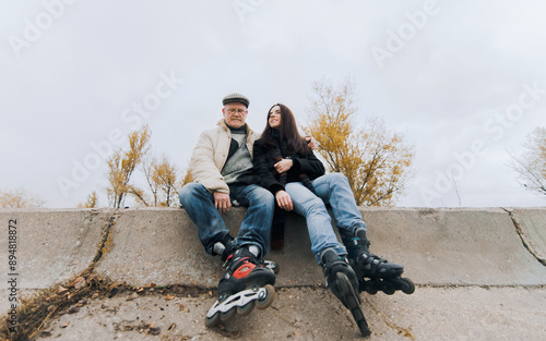 An elderly father in a down jacket, glasses, jeans and roller skates hugs his young daughter in a coat and roller skates. The concept of understanding between parents and children and happy retirement