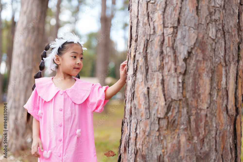 Asian girl in pink dress standing near tree in park. Looking to side, enjoying natural environment. Concept of childhood wonder, exploration, and playful moments in a sunlit forest setting. Asian girl in pink dress standing near tree in park. Looking to side, enjoying natural environment. Concept of childhood wonder, exploration, and playful moments in a sunlit forest setting.