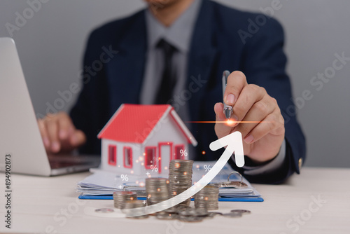 Businessman in suit analyzing increasing real estate market trends using a model house and stacked coins with an upward arrow and percentage symbols