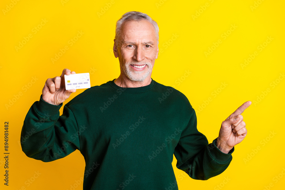 Photo portrait of handsome retired man hold credit card point empty space wear trendy green outfit isolated on yellow color background Photo portrait of handsome retired man hold credit card point empty space wear trendy green outfit isolated on yellow color background