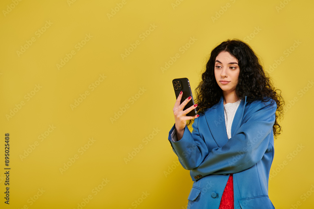 Naklejka premium Young brunette woman with curly hair, wearing a blue jacket, looking at her cell phone in a studio with a yellow background.