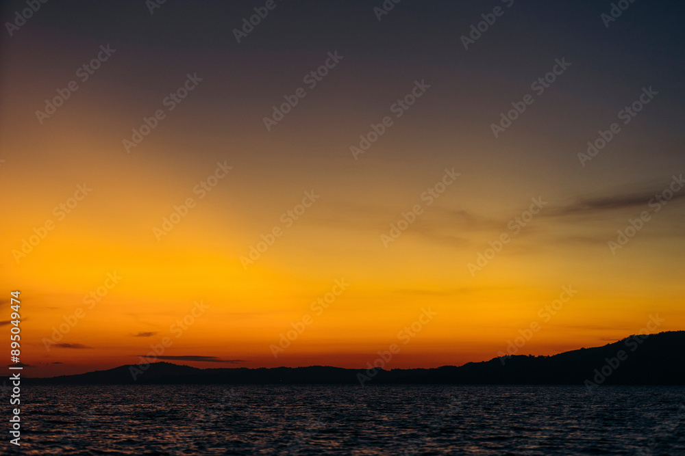 Warm glowing sunset light in the sky over the silhouetted island of Bunaken in the Celebes Sea, Indonesia; Bunaken, North Sulawesi, Indonesia Warm glowing sunset light in the sky over the silhouetted island of Bunaken in the Celebes Sea, Indonesia; Bunaken, North Sulawesi, Indonesia