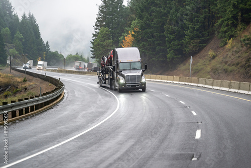 Gray bonnet big rig semi truck transporting cars on car hauler semi trailer driving on the winding highway road with rain and fog