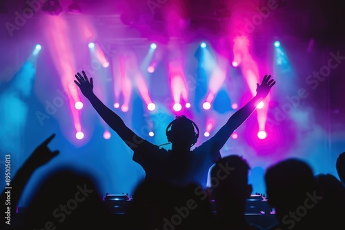 A DJ performs on stage at a lively music festival, bathed in vibrant pink and blue lights. The DJ's silhouette is visible against the bright stage lights, arms raised in excitement.