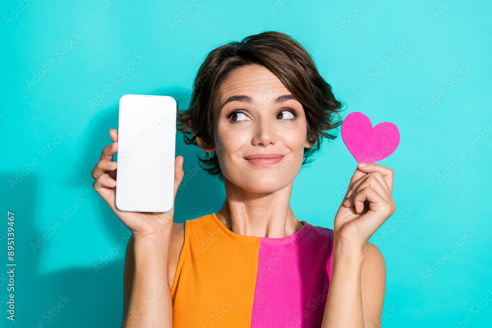 Photo of funky tricky lady dressed colorful top holding small heart looking modern gadget empty space isolated turquoise color background Photo of funky tricky lady dressed colorful top holding small heart looking modern gadget empty space isolated turquoise color background