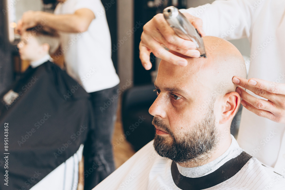 Fototapeta premium making hairstyle for man in a Barber shop, Latin American man gives a haircut to a bald bearded man in the barbershop salon