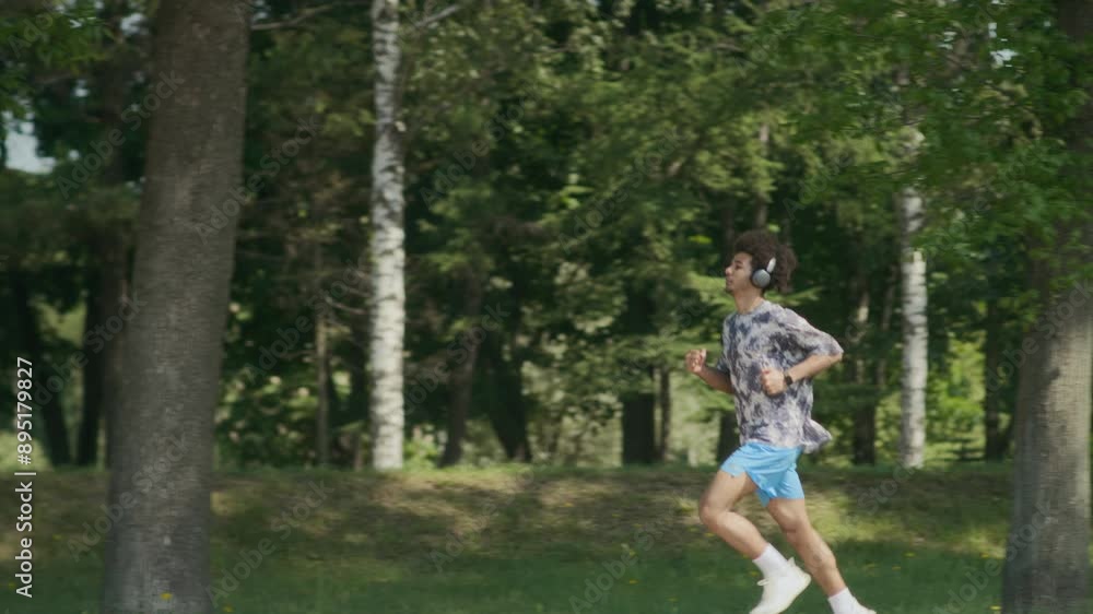 PAN slow motion shot of young man in headphones jogging through park while exercising outdoors on summer day PAN slow motion shot of young man in headphones jogging through park while exercising outdoors on summer day