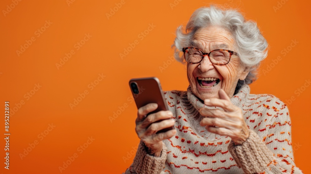 An elderly woman with gray hair is joyfully smiling and laughing while holding her phone against a vibrant orange background. An elderly woman with gray hair is joyfully smiling and laughing while holding her phone against a vibrant orange background.