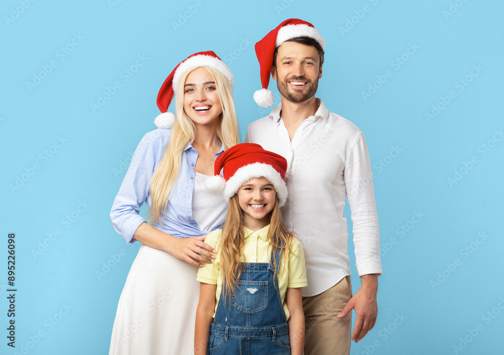 Obraz premium A young girl stands between her parents, all three smiling and wearing Santa hats. The family is standing in front of a solid blue background.