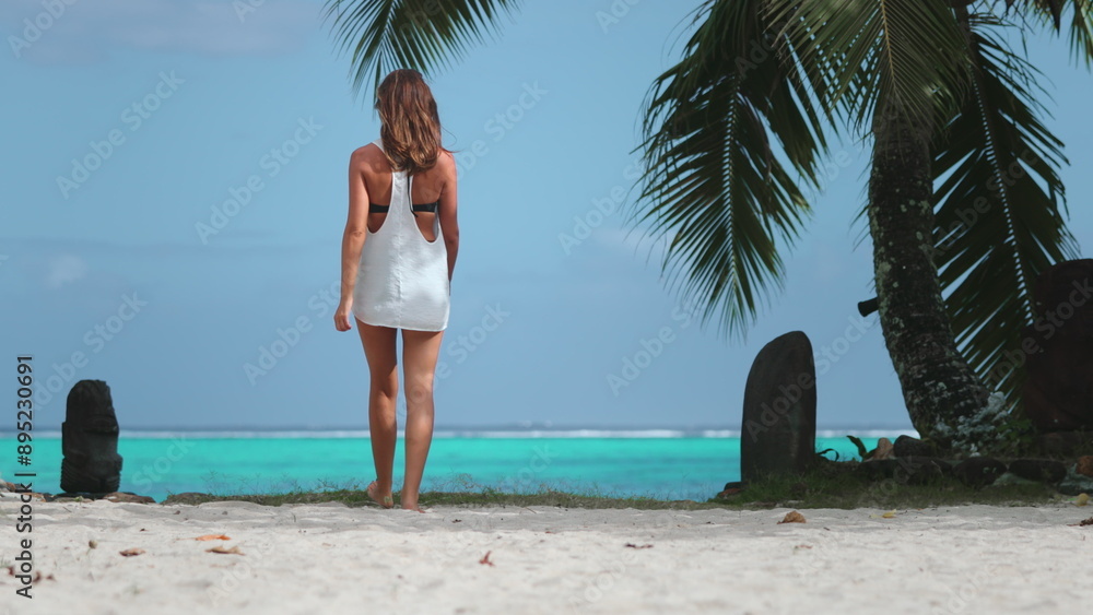 Obraz premium Woman in white dress walking on sandy beach, looking ocean. Travel, tourism, , rest relax concept. Palm tree, turquoise water. Outdoor lifestyle travel on summer holiday vacation. Back view