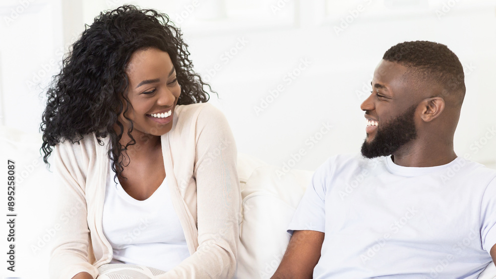 Obraz premium A happy couple sits together on a couch in a light-filled room. They are both smiling and looking at each other.