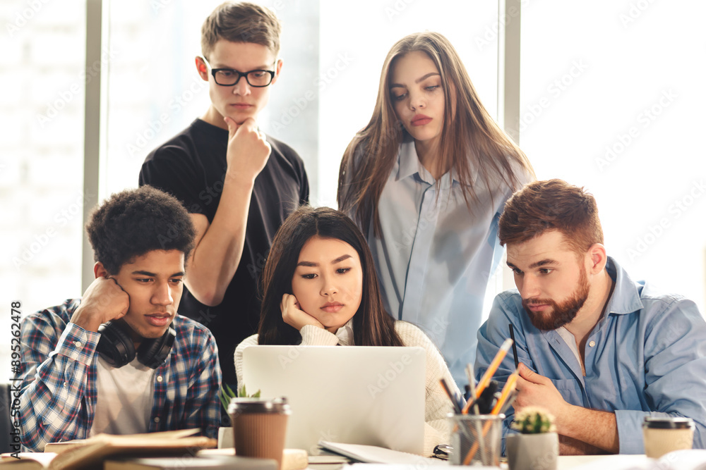 A group of five young professionals is gathered around a laptop, collaborating intently on a project. Some appearing pensive and others focused, they work together in a sunlit office setting A group of five young professionals is gathered around a laptop, collaborating intently on a project. Some appearing pensive and others focused, they work together in a sunlit office setting
