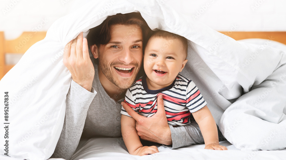 A father and his baby boy are lying under a white blanket in a bedroom. The father is smiling and looking at the camera, while the baby is looking up and laughing. A father and his baby boy are lying under a white blanket in a bedroom. The father is smiling and looking at the camera, while the baby is looking up and laughing.