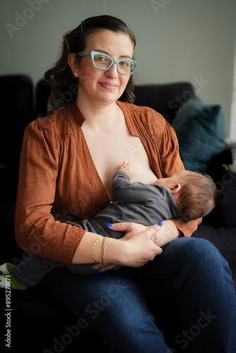 Young mother breastfeeding her baby, the woman is sitting on a sofa and smiling
