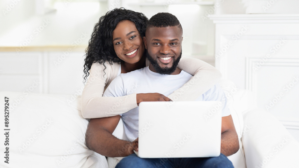 A couple sits together on a couch, smiling as they use a laptop. A couple sits together on a couch, smiling as they use a laptop.