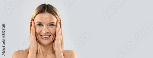 Photos Closeup of happy pretty nude middle aged blonde woman smiling at camera and touching face, posing on grey studio background, pulling face skin, copy space