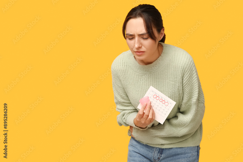 Young woman holding menstrual cup and calendar with marked days of menstruation on yellow background Young woman holding menstrual cup and calendar with marked days of menstruation on yellow background