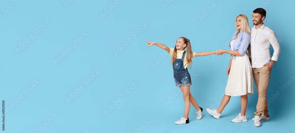 A young girl, a woman, and a man are walking and smiling, holding hands. The girl is pointing to copy space. They are all wearing casual clothing and standing against blue background.