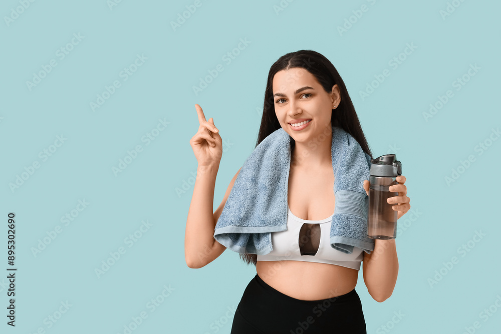 Beautiful young happy woman with towel and sports bottle of water pointing at something on blue background Beautiful young happy woman with towel and sports bottle of water pointing at something on blue background