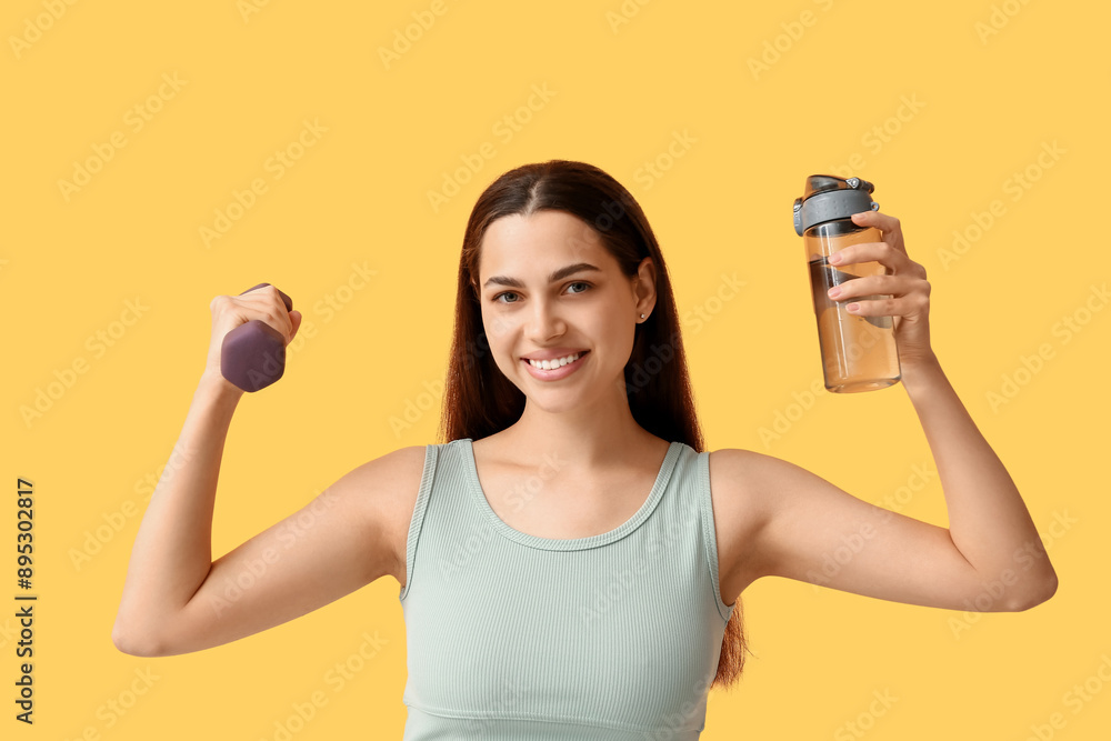 Beautiful young happy woman with dumbbell and sports bottle of water on yellow background