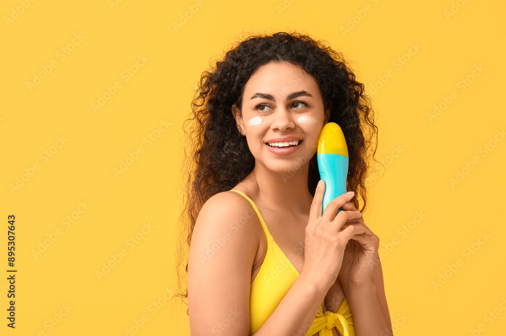 Beautiful young happy African-American woman with bottle of sunscreen cream on yellow background Beautiful young happy African-American woman with bottle of sunscreen cream on yellow background