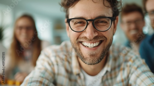 A cheerful man wearing glasses smiling brightly at the camera, with his friends out of focus in the background, creating a joyful and lively atmosphere.