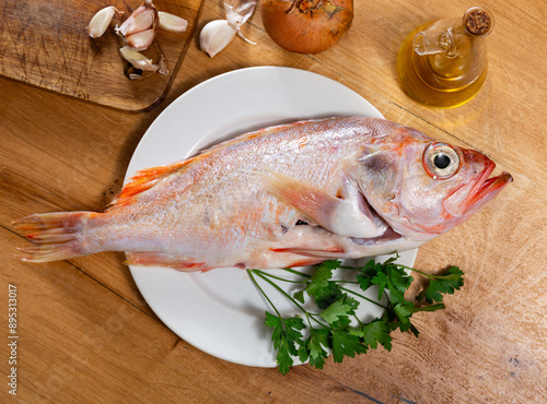 Raw longfinned beryx with fresh greens, spicy garlic, onion and decanter of olive oil arranged on white plate on wooden background. Cooking ingredients.. photo