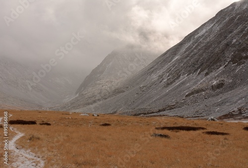 A narrow winding path crosses a valley with yellowed short grass in a crevice at the junction of two mountain ranges on an overcast autumn day.