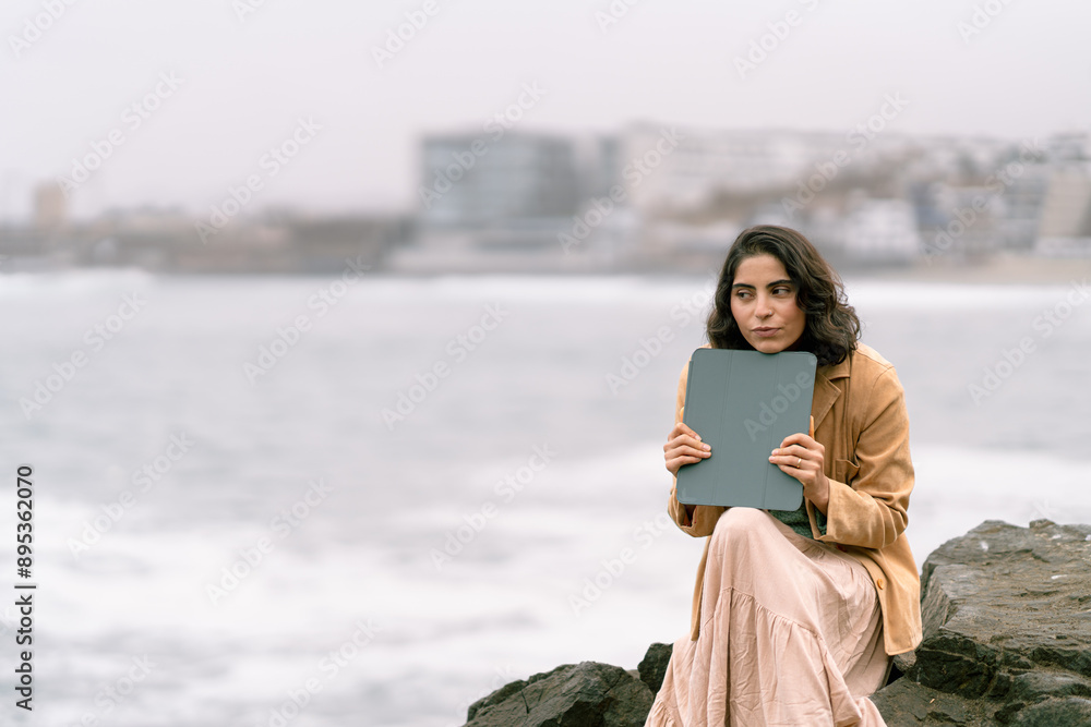 Thoughtful woman holding a tablet and looking at the ocean on a winter day Thoughtful woman holding a tablet and looking at the ocean on a winter day