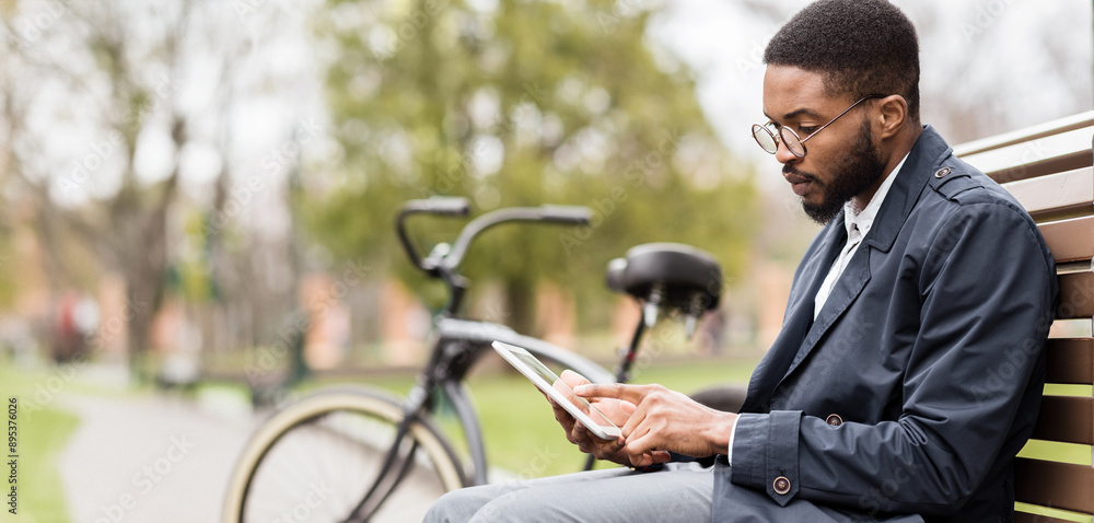 An African American man is on a park bench, working on a digital tablet with a bike nearby. The setting is calm and green, reflecting a mix of business and leisure in nature. An African American man is on a park bench, working on a digital tablet with a bike nearby. The setting is calm and green, reflecting a mix of business and leisure in nature.