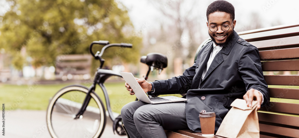 Obraz premium A professional black man uses his laptop on a park bench with a bicycle nearby. The atmosphere is serene and efficient, capturing the essence of outdoor productivity.