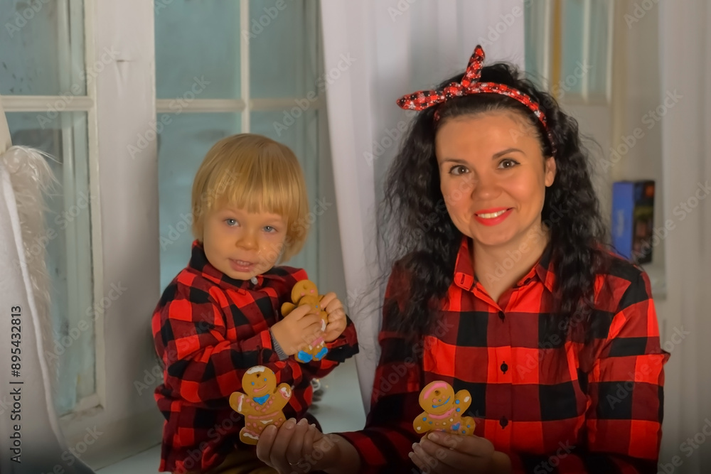 Mom and son with christmas cookies by the window. Mother and son dressed in identical festive Christmas clothes in a check sitting on the windowsill with a Christmas cookies. Family look. Christmas Mom and son with christmas cookies by the window. Mother and son dressed in identical festive Christmas clothes in a check sitting on the windowsill with a Christmas cookies. Family look. Christmas