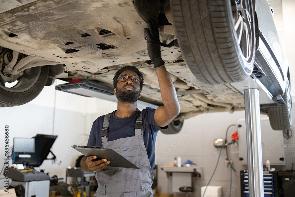 Mechanic inspecting car on lift in auto repair shop. Professional technician examining underside of vehicle while holding clipboard. Routine maintenance and safety check in modern garage. Mechanic inspecting car on lift in auto repair shop. Professional technician examining underside of vehicle while holding clipboard. Routine maintenance and safety check in modern garage.