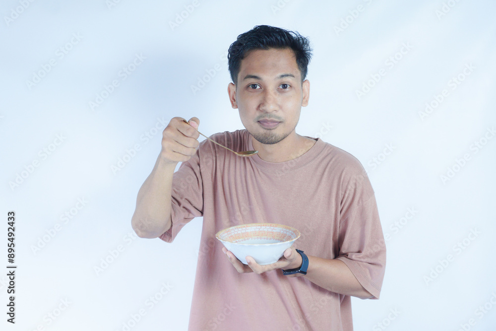 A portrait of an Indonesian Asian man wearing a pink tshirt, seen happily posing and enjoying his meal with a spoon and bowl, isolated on a green background.