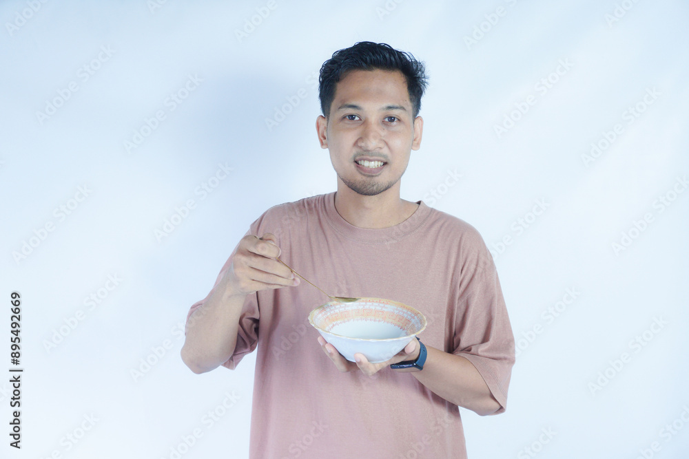 A portrait of an Indonesian Asian man wearing a pink tshirt, seen happily posing and enjoying his meal with a spoon and bowl, isolated on a green background. A portrait of an Indonesian Asian man wearing a pink tshirt, seen happily posing and enjoying his meal with a spoon and bowl, isolated on a green background.