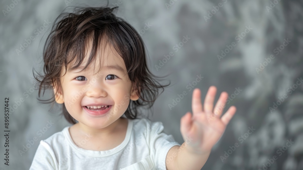 Adorable smiling toddler waving cheerfully in front of a grey background, wearing a white shirt, engaging and happy expression. Adorable smiling toddler waving cheerfully in front of a grey background, wearing a white shirt, engaging and happy expression.