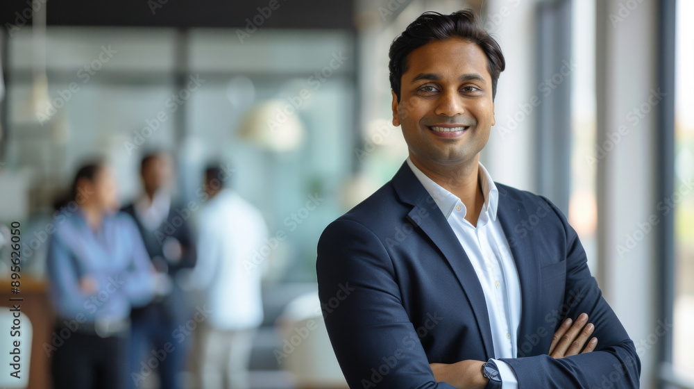 A confident young professional stands with arms crossed, smiling warmly inside a bright modern office with colleagues in the background. A confident young professional stands with arms crossed, smiling warmly inside a bright modern office with colleagues in the background.