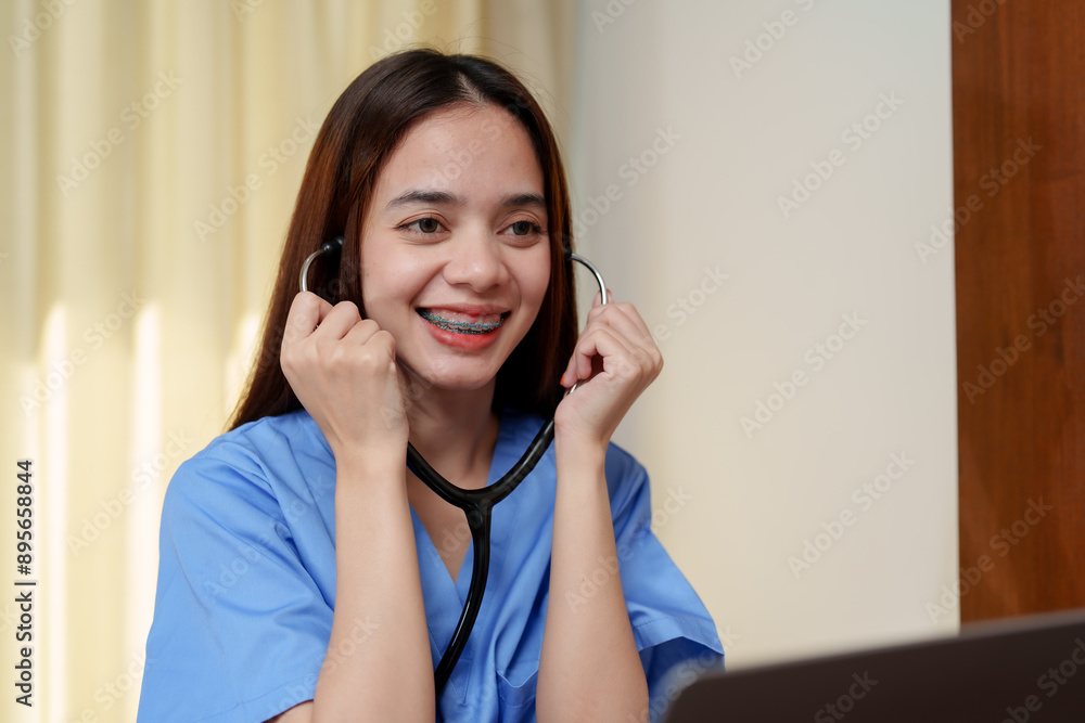 Smiling Asian teenage girl in blue medical scrubs, holding stethoscope to ears, sitting at desk with laptop. Warm lighting and curtain background. Emphasizes happiness, medical study, and preparation. Smiling Asian teenage girl in blue medical scrubs, holding stethoscope to ears, sitting at desk with laptop. Warm lighting and curtain background. Emphasizes happiness, medical study, and preparation.