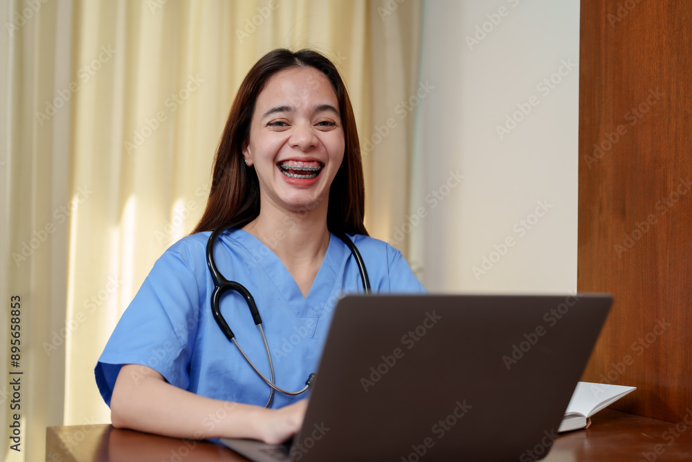 Asian teenage girl with long hair, wearing blue medical scrubs and a stethoscope, smiling and working on a laptop. Light coming through curtains in the background, creating a warm atmosphere. Asian teenage girl with long hair, wearing blue medical scrubs and a stethoscope, smiling and working on a laptop. Light coming through curtains in the background, creating a warm atmosphere.