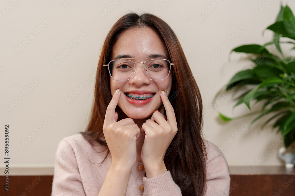 Asian teenage girl with braces, smiling and pointing at her cheeks, emphasizing her smile. Wearing glasses, indoors with soft lighting. Represents joy, self-confidence, and happiness on rest days. Asian teenage girl with braces, smiling and pointing at her cheeks, emphasizing her smile. Wearing glasses, indoors with soft lighting. Represents joy, self-confidence, and happiness on rest days.
