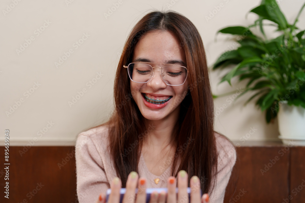 Asian teenage girl with braces, smiling and wearing glasses, looking at her nails. Wearing pink sweater, indoors with soft lighting. Represents self-care, happiness, and casual relaxation on rest days Asian teenage girl with braces, smiling and wearing glasses, looking at her nails. Wearing pink sweater, indoors with soft lighting. Represents self-care, happiness, and casual relaxation on rest days