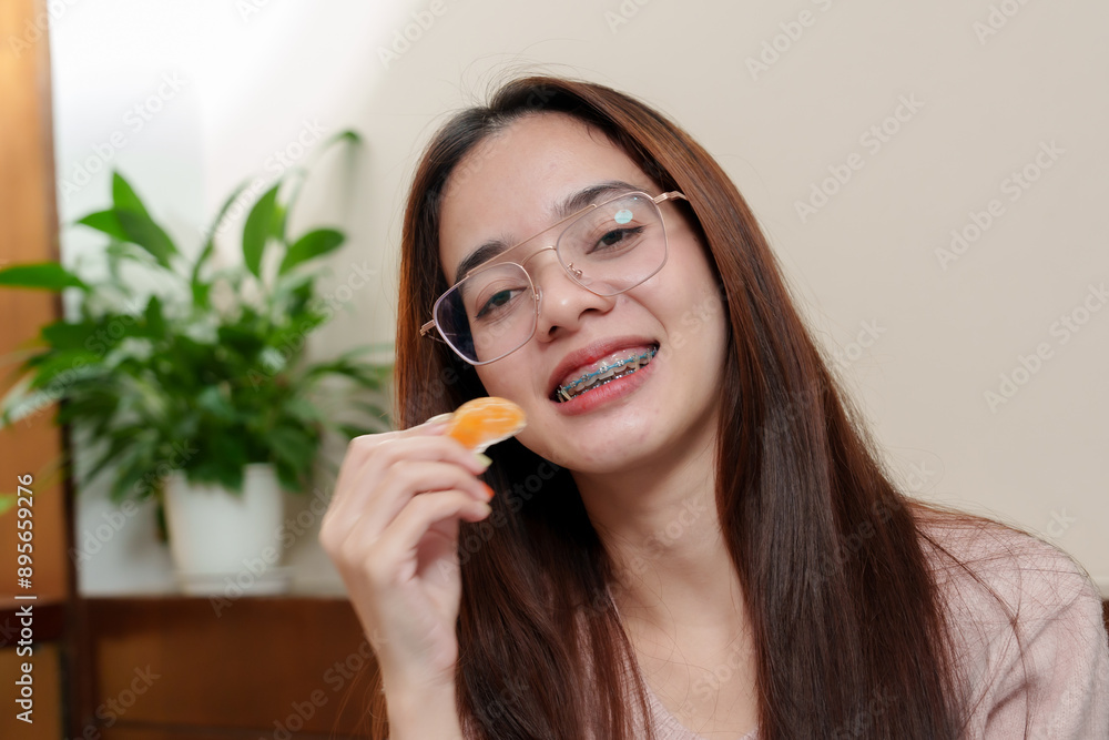 Asian teenage girl with braces, smiling and holding an orange slice near her face. Wearing glasses and pink sweater, indoors with soft lighting. healthy eating, and relaxation on rest days. Asian teenage girl with braces, smiling and holding an orange slice near her face. Wearing glasses and pink sweater, indoors with soft lighting. healthy eating, and relaxation on rest days.